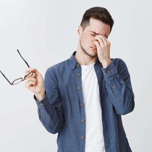 Guy rubbing eyes as feeling tired after wearing glasses while working over freelance project on computer standing with eyewear in hand and closed eyes being drained and exhausted, being hardworking. Copy space